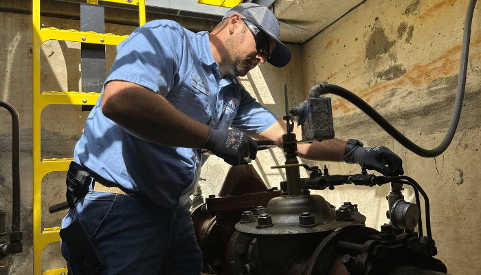 A worker in a blue shirt and safety gloves repairs an engine component inside a workshop, using tools while leaning under a yellow ladder and beside a concrete wall with stains